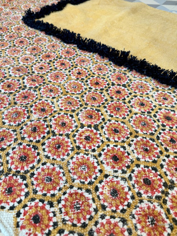 A close-up of a woven textile with a repeating floral or medallion pattern in red, orange, white, black, and yellow, with a mustard yellow section and black fringe visible at the top.