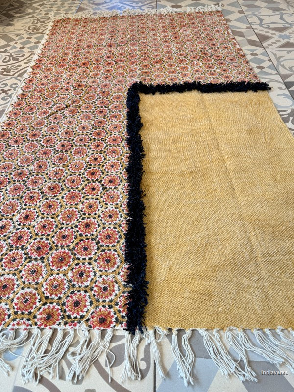 A rectangular woven rug laid flat on a tiled floor, showing two sides: one side has a dense multicolor floral and geometric pattern in pink, orange, green, and black on a light base, and the other side is a plain warm beige with a black fringed border. White fringe is visible along the bottom edge.