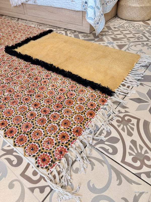 A rectangular area rug shown on a patterned tile floor in a bedroom setting, with part of a bed and a woven basket visible. The rug has a mustard yellow top section with a black fringe, a cream section with white fringe, and a floral patterned section in rust red, black, and cream.