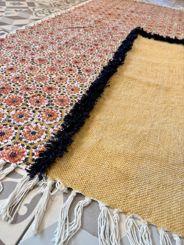 A close-up of a woven rug laid on a tiled floor, showing a mustard yellow side with white fringe and a patterned side with pink, red, black, and white floral motifs.