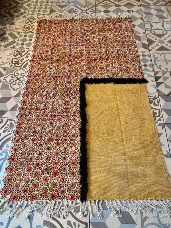 A folded and unfolded view of a traditional Moroccan wool rug on a patterned tile floor. The rug features a dense red, black, and beige floral pattern on one half, and a solid golden-yellow side with black fringe and white tassels.