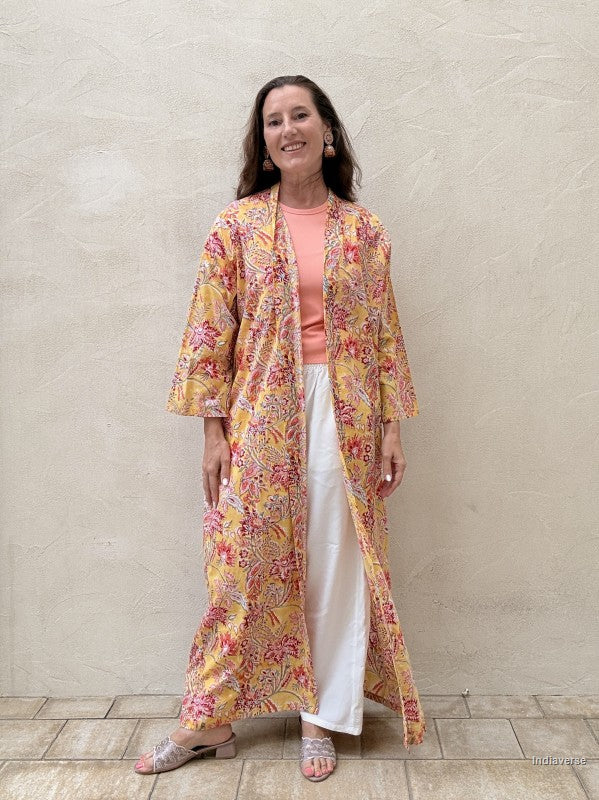 Woman standing n front of a beige wall wearing a long floral robe in yellow with red flowers