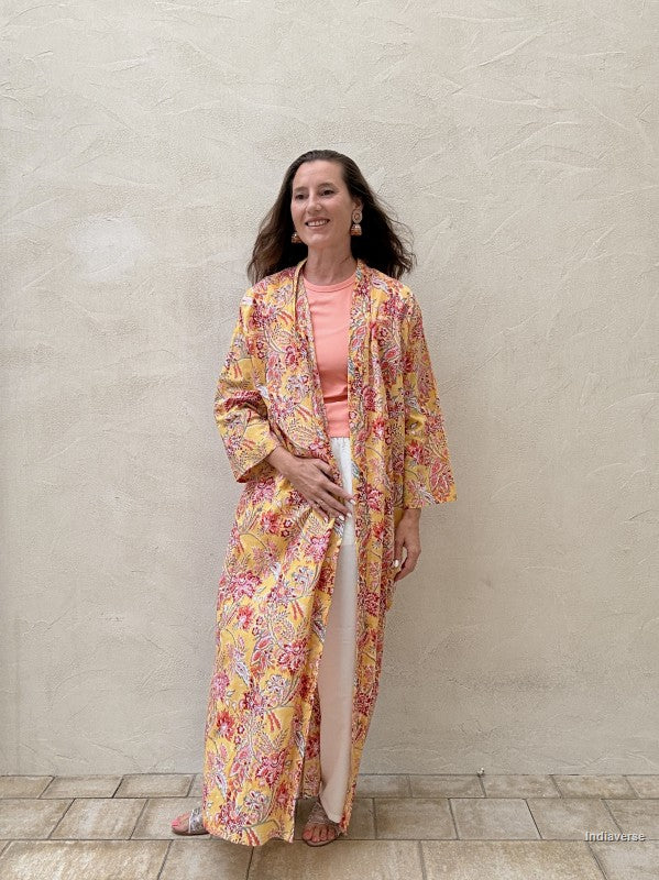 Woman standing n front of a beige wall wearing a long floral robe in yellow with red flowers