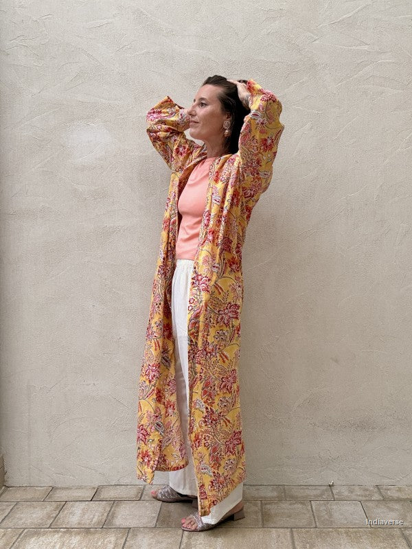 Woman standing n front of a beige wall wearing a long floral robe in yellow with red flowers