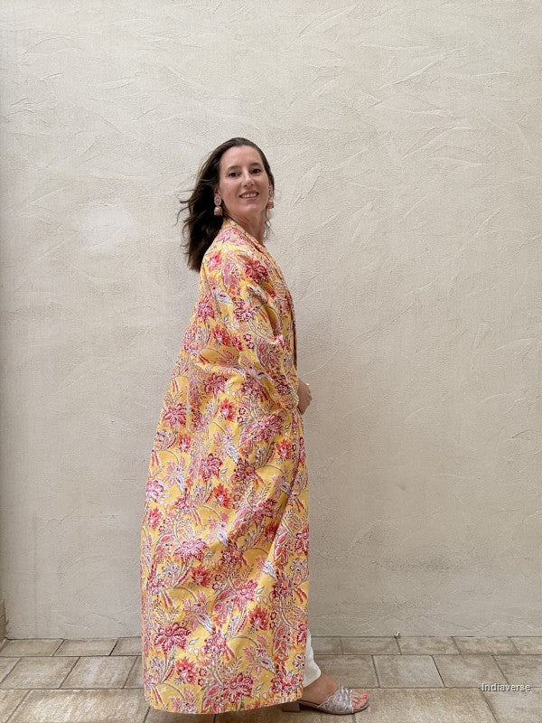 Woman standing n front of a beige wall wearing a long floral robe in yellow with red flowers
