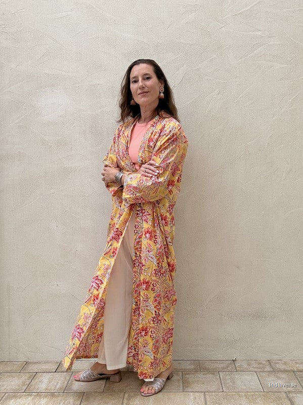 Woman standing n front of a beige wall wearing a long floral robe in yellow with red flowers