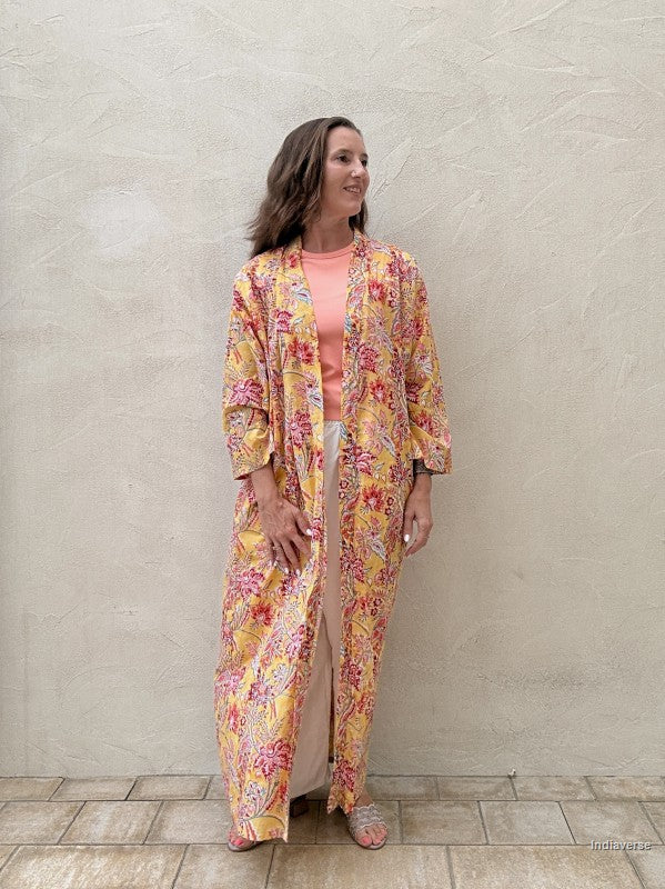 Woman standing n front of a beige wall wearing a long floral robe in yellow with red flowers