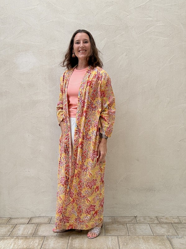 Woman standing n front of a beige wall wearing a long floral robe in yellow with red flowers