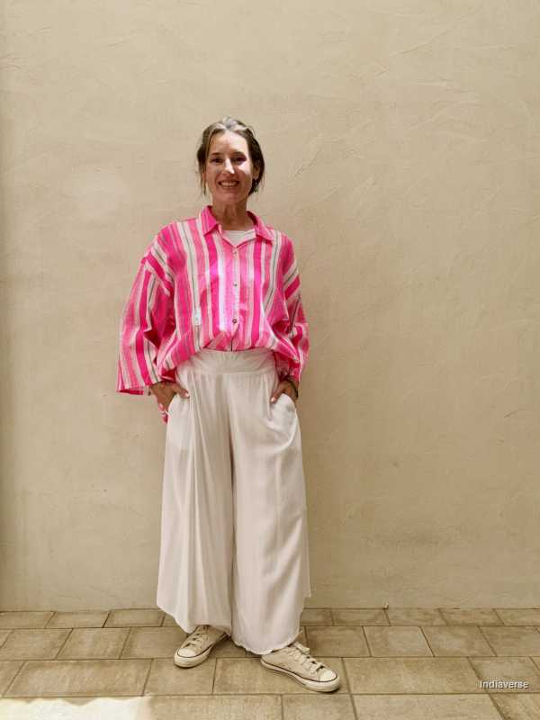 Person wearing a pink striped shirt and white pants standing against a beige wall.