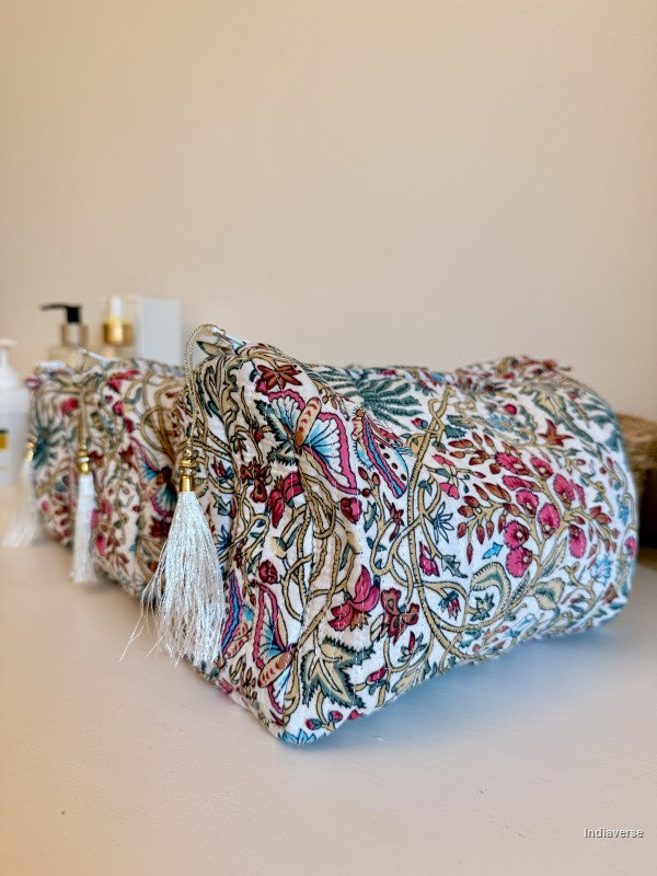 A cylindrical cosmetic pouch with a dense floral and paisley print in pink, blue, green, and gold on a white base, featuring white tassels with gold bead accents, resting on a beige surface | Palm Paradise