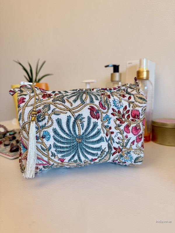 A small zippered cosmetic pouch with an intricate multicolor floral and paisley print on a white base, featuring a white tassel charm, displayed on a beige tabletop with a neutral background | Palm Paradise