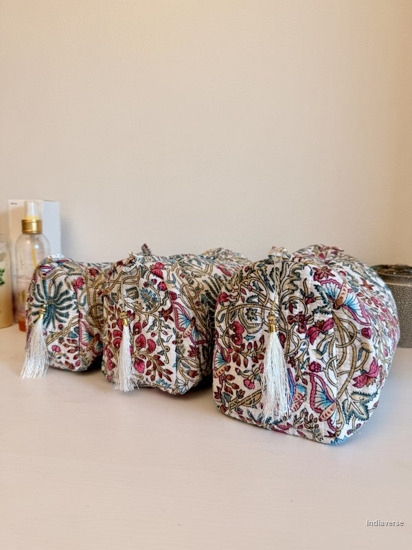Three small drawstring cosmetic pouches with white tassels, featuring a dense multicolor floral and paisley block print on a cream fabric base, arranged on a light beige surface against a neutral beige background | Palm Paradise