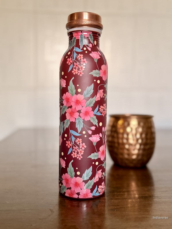 copper water bottle with red and pink floral design and a Bordeaux color background sitting on a brown table with a copper cup in the background