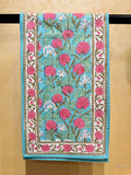 Beautiful table runner with a hand-block printed floral pattern. The design features vibrant pink and white flowers with green stems on a turquoise blue background, framed by a white border with pink floral motifs.