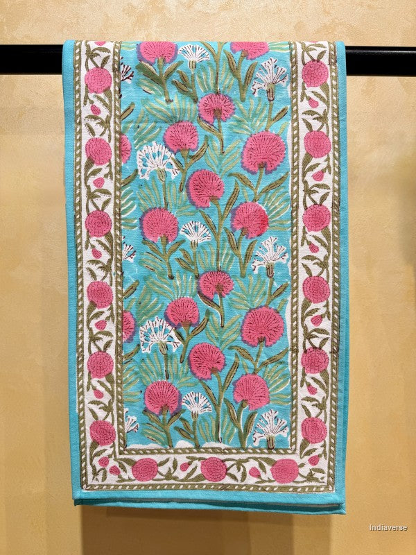 Beautiful table runner with a hand-block printed floral pattern. The design features vibrant pink and white flowers with green stems on a turquoise blue background, framed by a white border with pink floral motifs.