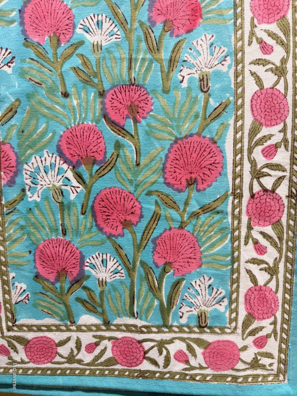 Table runner with a hand-block printed floral pattern. The design features vibrant pink and white flowers with green stems on a turquoise blue background, framed by a white border with pink floral motifs.