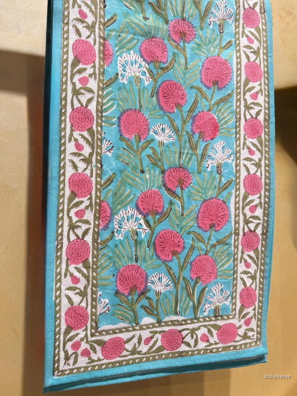 A close-up of a textile or fabric panel with a hand-block printed floral pattern. The design features vibrant pink and white flowers with green stems on a turquoise blue background, framed by a white border with pink floral motifs.