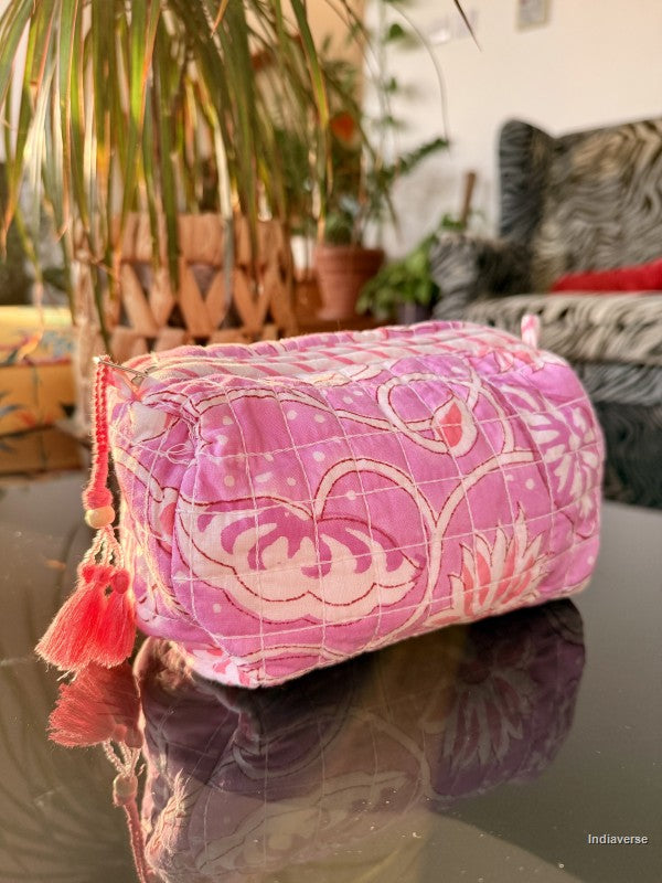 A pink quilted fabric cosmetic pouch with a white floral pattern and red tassels, resting on a reflective dark surface with a blurred indoor background.