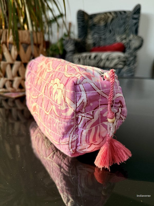 A pink quilted cosmetic pouch with a floral pattern and a matching tassel zipper pull, resting on a dark reflective surface with a blurred indoor background.