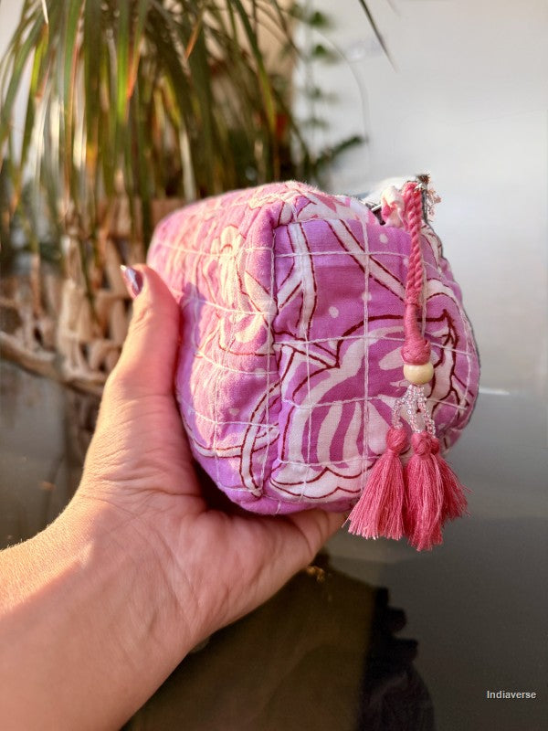 A hand holding a small pink quilted pouch with a white floral print and a pink tassel zipper pull, set against a blurred background of a plant and neutral wall.