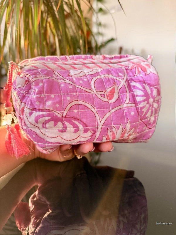 A pink quilted fabric cosmetic pouch with white floral patterns and pink tassels, held by a hand in front of a softly blurred indoor background with warm sunlight.