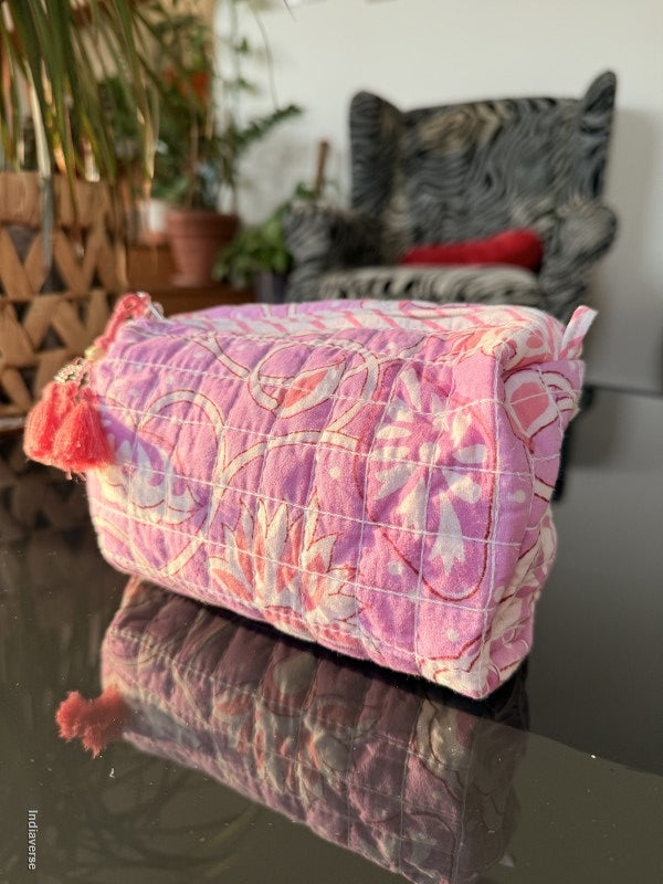 A pink quilted cushion cover with a floral and paisley pattern, shown in a home setting next to another cushion cover on a glass table.