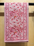 A pink embroidered cotton textile, likely a handkerchief or small towel, hanging from a black rod against a textured beige wall. The fabric features intricate floral embroidery in shades of pink, red, and brown.
