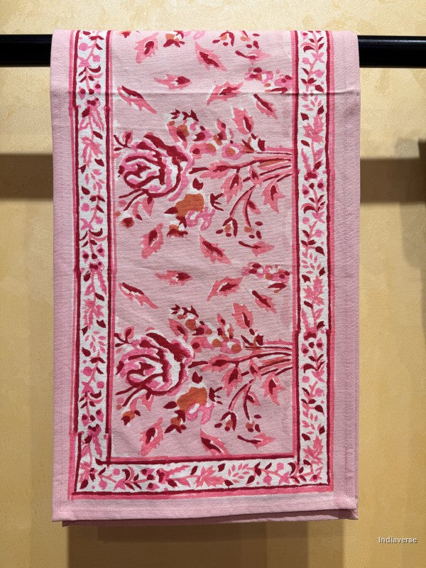A pink embroidered cotton textile, likely a handkerchief or small towel, hanging from a black rod against a textured beige wall. The fabric features intricate floral embroidery in shades of pink, red, and brown.