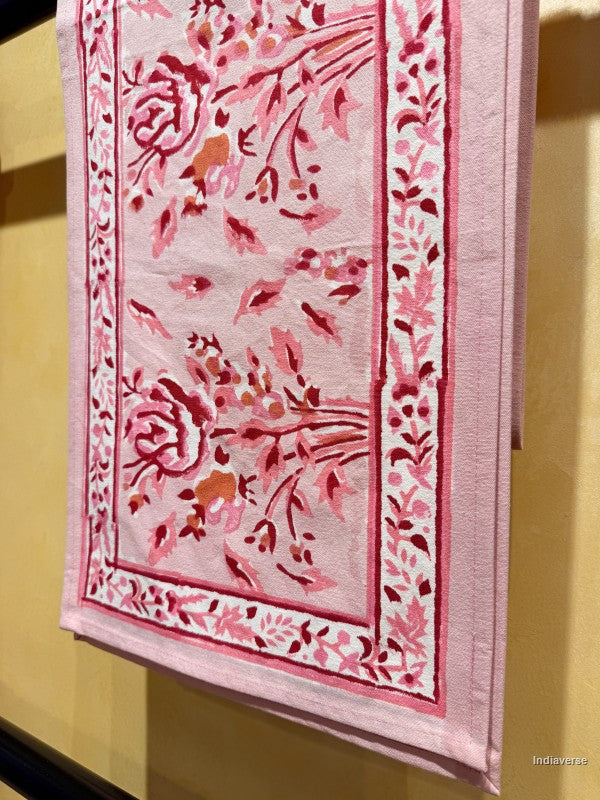A pink cotton towel featuring a floral and botanical pattern in shades of red, rose, and orange, hanging against a warm beige wall.