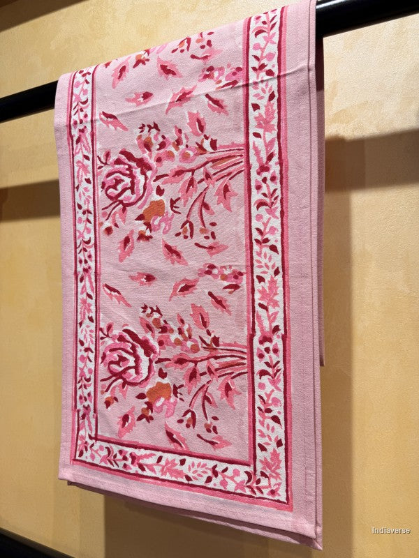 A pink cotton kitchen towel with a floral and botanical pattern, hanging from a black rod against a beige wall.