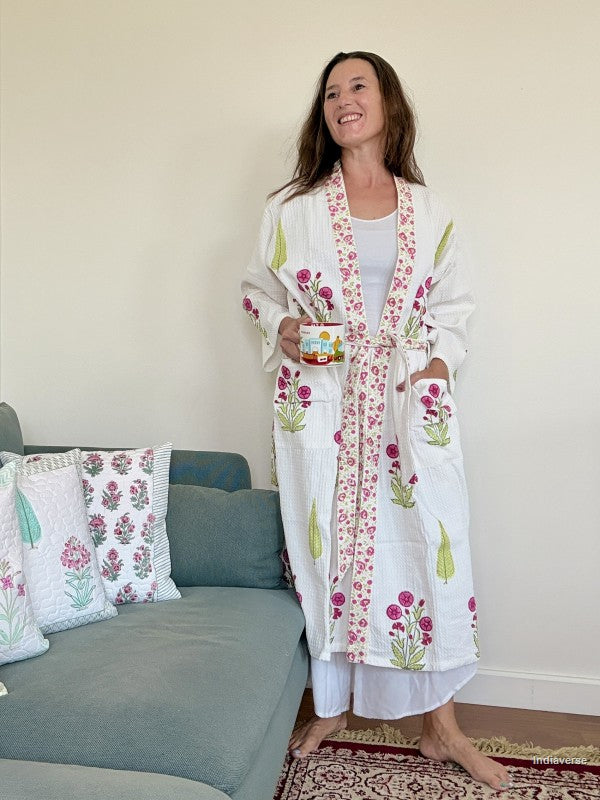 A woman wearing a white cotton robe with pink and green floral embroidery stands in a living room setting, holding a mug next to a blue-gray sofa with matching embroidered pillows.