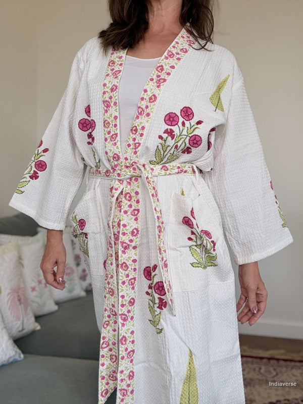 A woman wearing a white cotton waffle-weave kimono-style robe with pink floral embroidery and green leaf accents, tied at the waist. The background shows a softly blurred indoor setting with a sofa and pillows.