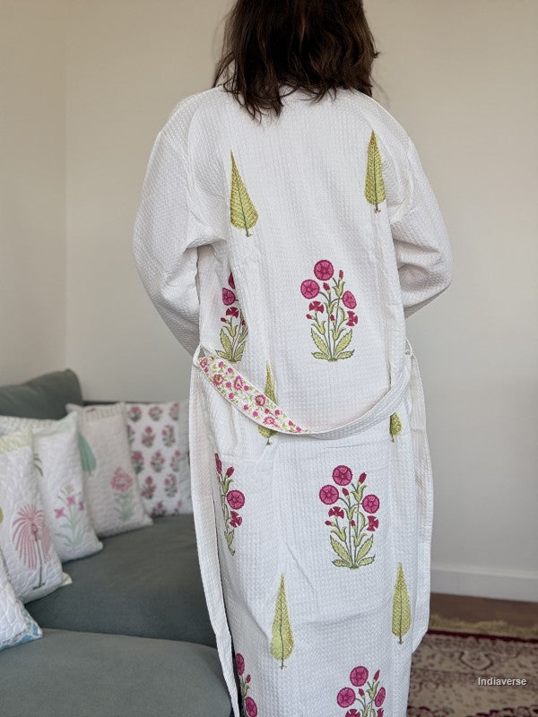 A person viewed from behind wearing a white waffle-knit robe with pink floral and green tree prints, standing in a living room with a sofa and pillows in the background.