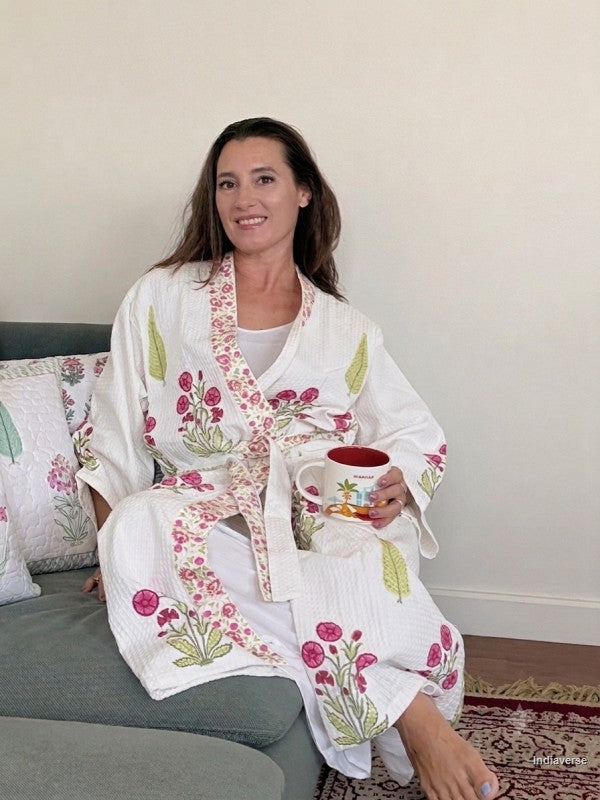 A woman sitting on a gray sofa wearing a white waffle-knit robe with pink floral and green leaf patterns, holding a mug.
