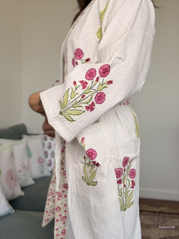 A person wearing a white textured waffle-knit robe with pink floral embroidery and green leaves, standing indoors near a sofa with patterned pillows.