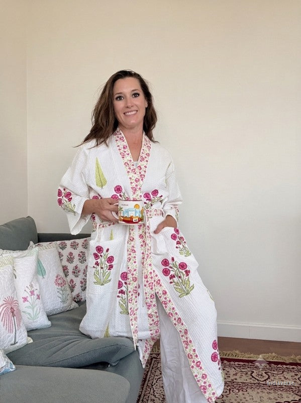 A woman wearing a white cotton kimono-style robe with pink floral and green leaf embroidery, standing in a living room and holding a small colorful mug.