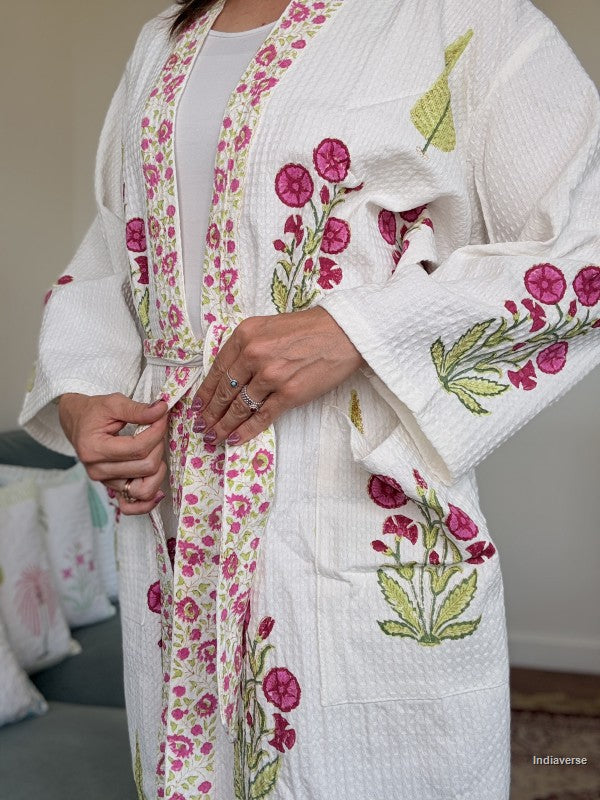 A person wearing a white textured kimono-style robe with pink floral embroidery and green leaf details, shown from the shoulders to the knees in an indoor setting.