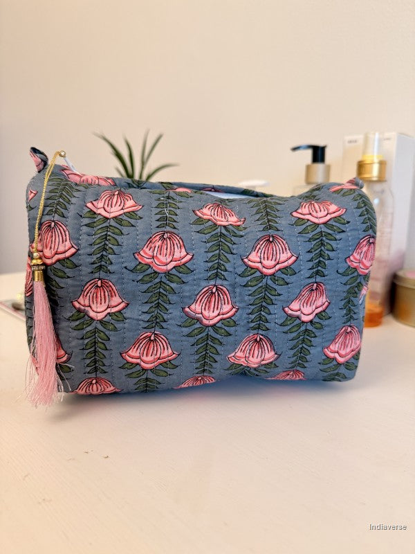 A fabric cosmetic pouch with a blue base, pink bell-shaped floral pattern, and a pink tassel zipper pull, shown on a tabletop with toiletry bottles and a plant in the background | Pink Lotus