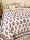 A floral quilted bedspread with matching pillowcases, featuring a pink and green floral pattern on a white background with a decorative red and green border, styled on a bed.