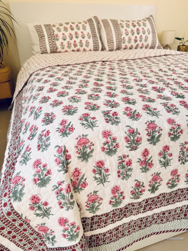 A floral quilted bedspread with matching pillowcases, featuring a pink and green floral pattern on a white background with a decorative red and green border, styled on a bed.