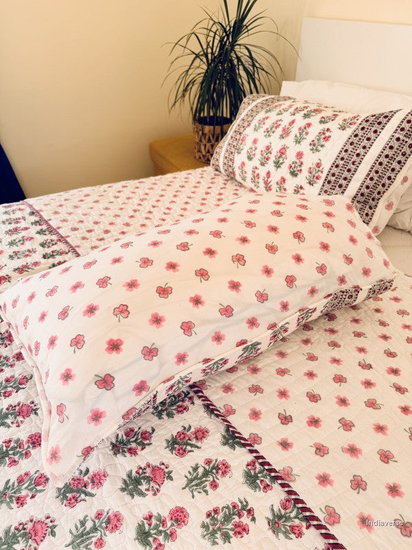 A floral quilted bedspread with matching pillowcases, featuring a pink and green floral pattern on a white background with a decorative red and green border, styled on a bed.