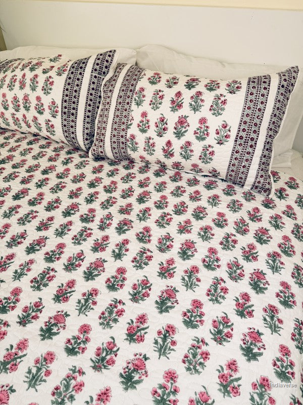 A floral quilted bedspread with matching pillowcases, featuring a pink and green floral pattern on a white background with a decorative red and green border, styled on a bed.