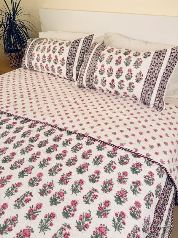 A floral quilted bedspread with matching pillowcases, featuring a pink and green floral pattern on a white background with a decorative red and green border, styled on a bed.