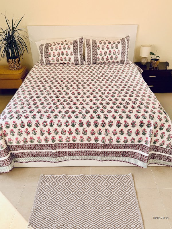 A floral quilted bedspread with matching pillowcases, featuring a pink and green floral pattern on a white background with a decorative red and green border, styled on a bed.