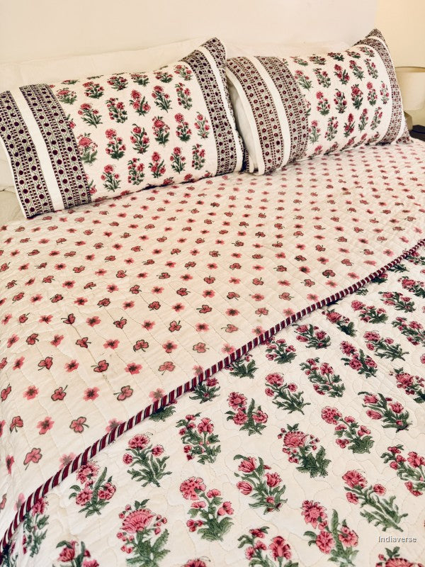 Close up of a floral quilted bedspread with matching pillowcases, featuring a pink and green floral pattern on a white background with a decorative red and green border, styled on a bed.
