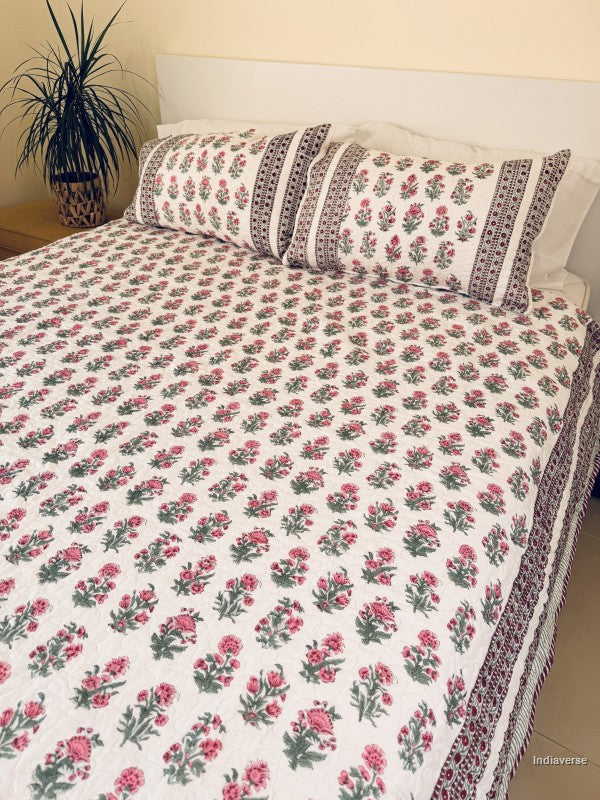 A floral quilted bedspread with matching pillowcases, featuring a pink and green floral pattern on a white background with a decorative red and green border, styled on a bed.