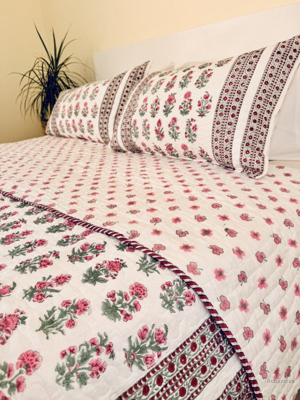 A floral quilted bedspread with matching pillowcases, featuring a pink and green floral pattern on a white background with a decorative red and green border, styled on a bed.