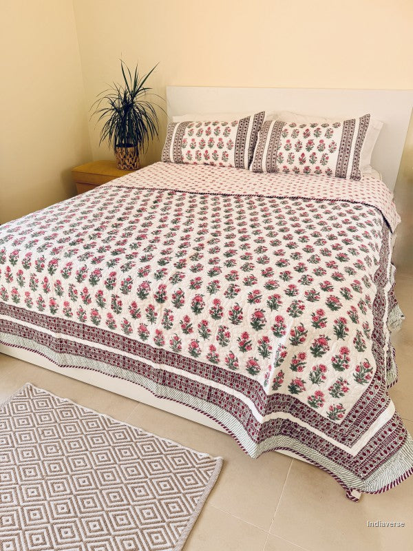 A floral quilted bedspread with matching pillowcases, featuring a pink and green floral pattern on a white background with a decorative red and green border, styled on a bed.