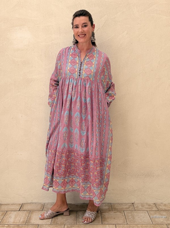 A woman wearing a long-sleeved, loose-fitting bohemian-style maxi dress with a pink, blue, and orange paisley pattern, standing against a textured beige stucco wall.