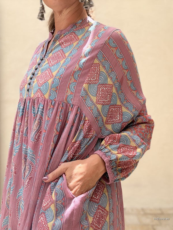 A close-up of a woman wearing a dusty pink long-sleeve tunic dress with a colorful geometric and paisley print in blue, yellow, and red, set against a neutral beige background.
